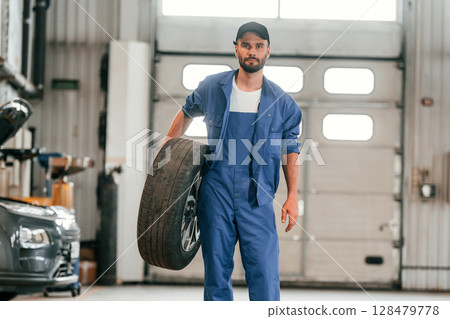Front view, standing and holding tire. Auto mechanic working in garage. Repair service Front view, standing and holding tire. Auto mechanic working in garage. Repair service 128479778