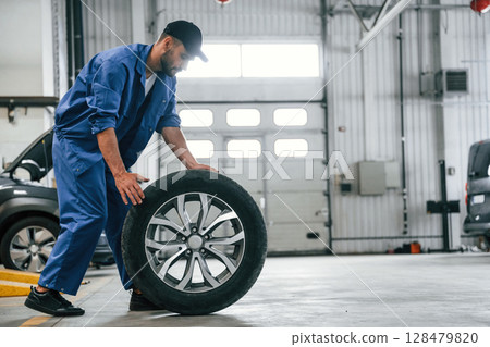New tire, moving it. Auto mechanic working in garage. Repair service New tire, moving it. Auto mechanic working in garage. Repair service 128479820