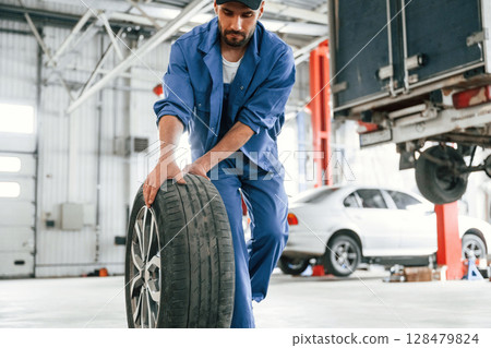 Walking forward with new tire. Auto mechanic working in garage. Repair service Walking forward with new tire. Auto mechanic working in garage. Repair service 128479824