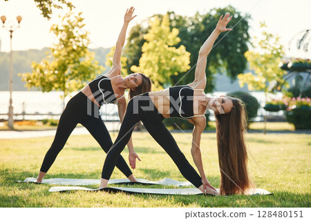 Doing yoga exercises. Two women in sport clothes are doing exercises outdoors on the field Doing yoga exercises. Two women in sport clothes are doing exercises outdoors on the field 128480151