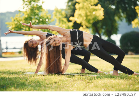 On the yoga mat. Two women in sport clothes are doing exercises outdoors on the field 128480157
