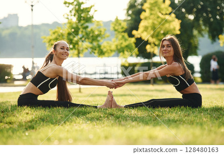 Holding each other by hands, touching feet. Two women in sport clothes are doing exercises outdoors on the field Holding each other by hands, touching feet. Two women in sport clothes are doing exercises outdoors on the field 128480163