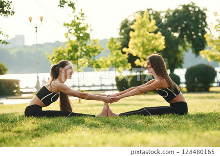 Holding each other by hands, touching feet. Two women in sport clothes are doing exercises outdoors on the field 128480165