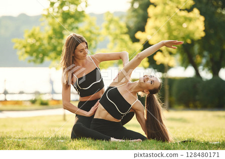 Professional trainer is helping. Two women in sport clothes are doing exercises outdoors on the field 128480171