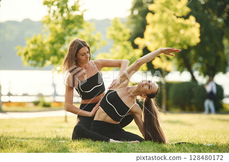 Professional trainer is helping. Two women in sport clothes are doing exercises outdoors on the field Professional trainer is helping. Two women in sport clothes are doing exercises outdoors on the field 128480172