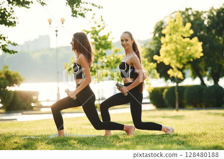 Dumbbells in hands. Two women in sport clothes are doing exercises outdoors on the field Dumbbells in hands. Two women in sport clothes are doing exercises outdoors on the field 128480188