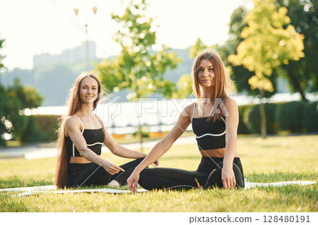Looking to the camera. Two women in sport clothes are doing exercises outdoors on the field Looking to the camera. Two women in sport clothes are doing exercises outdoors on the field 128480191