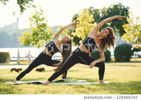 Asana named warrior in yoga. Two women in sport clothes are doing exercises outdoors on the field 128480192