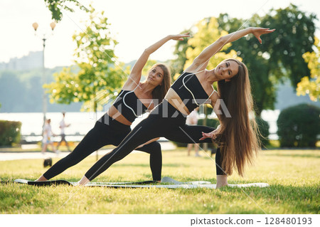Asana named warrior in yoga. Two women in sport clothes are doing exercises outdoors on the field Asana named warrior in yoga. Two women in sport clothes are doing exercises outdoors on the field 128480193