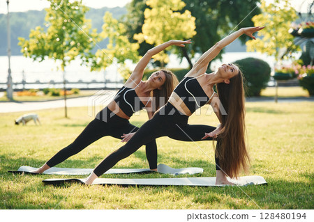 Asana named warrior in yoga. Two women in sport clothes are doing exercises outdoors on the field 128480194