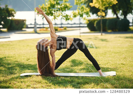 On the yoga mat. Beautiful young woman in sportive clothes is in the park at sunny daytime 128480329