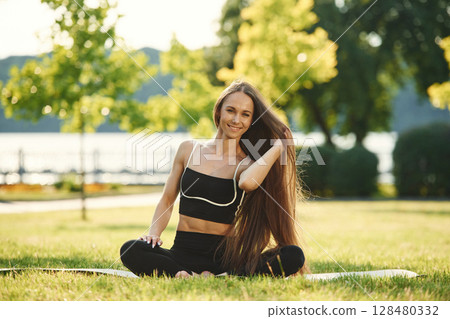Sitting on the yoga mat. Beautiful young woman in sportive clothes is in the park at sunny daytime Sitting on the yoga mat. Beautiful young woman in sportive clothes is in the park at sunny daytime 128480332