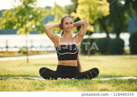 Sitting on the yoga mat. Beautiful young woman in sportive clothes is in the park at sunny daytime 128480334