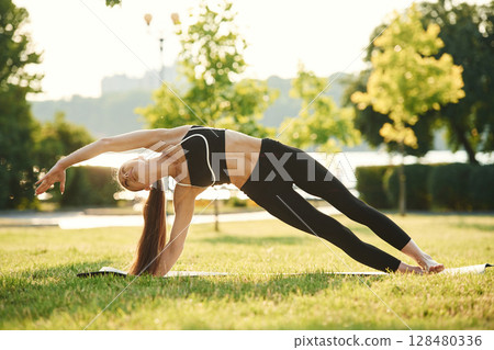 Exercises for flexibility. Beautiful young woman in sportive clothes is in the park at sunny daytime 128480336