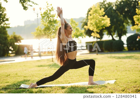 Doing squats. Beautiful young woman in sportive clothes is in the park at sunny daytime 128480341