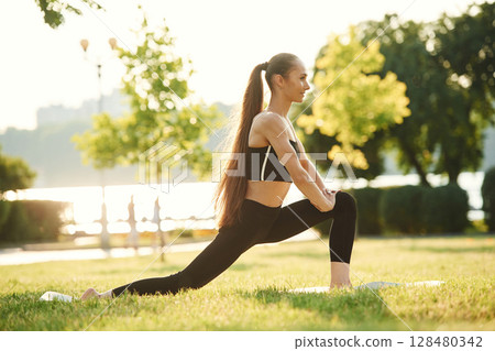 Doing squats. Beautiful young woman in sportive clothes is in the park at sunny daytime 128480342