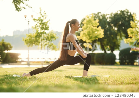 Doing squats. Beautiful young woman in sportive clothes is in the park at sunny daytime 128480343