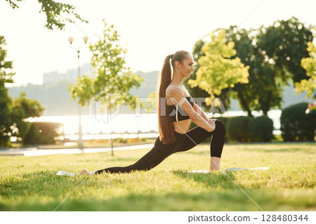 Doing squats. Beautiful young woman in sportive clothes is in the park at sunny daytime 128480344