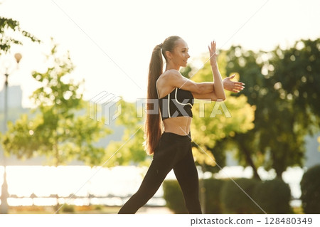 Preparing for the fitness session. Beautiful young woman in sportive clothes is in the park at sunny daytime 128480349