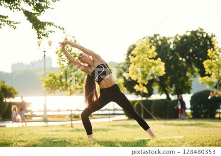 Standing, stretching the hands. Beautiful young woman in sportive clothes is in the park at sunny daytime 128480353