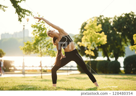 Standing, stretching the hands. Beautiful young woman in sportive clothes is in the park at sunny daytime 128480354