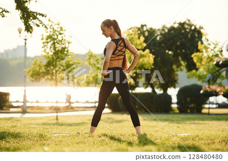 Hands on the waist, standing, rear view. Beautiful young woman in sportive clothes is in the park at sunny daytime 128480480