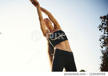 Stretches upwards, against blue sky. Beautiful young woman in sportive clothes is in the park at sunny daytime 128480498