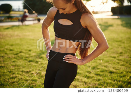 Showing abs. Beautiful young woman in sportive clothes is in the park at sunny daytime Showing abs. Beautiful young woman in sportive clothes is in the park at sunny daytime 128480499