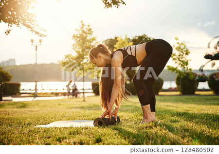 Rolling on yoga mat. Beautiful young woman in sportive clothes is in the park at sunny daytime Rolling on yoga mat. Beautiful young woman in sportive clothes is in the park at sunny daytime 128480502