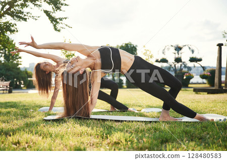 Warrior yoga asana, exercise. Two women in sport clothes are doing exercises outdoors on the field Warrior yoga asana, exercise. Two women in sport clothes are doing exercises outdoors on the field 128480583