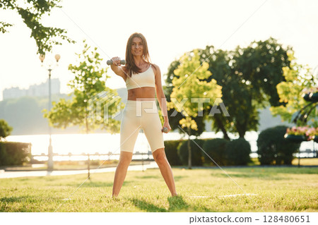 Holding dumbbell. Beautiful young woman in sportive clothes is in the park at sunny daytime Holding dumbbell. Beautiful young woman in sportive clothes is in the park at sunny daytime 128480651