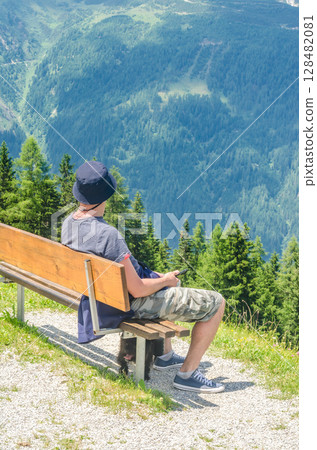 Man Relaxing on Bench Overlooking Forested Alpine Valley with Dog Resting Below. High quality photo Man Relaxing on Bench Overlooking Forested Alpine Valley with Dog Resting Below. High quality photo 128482081