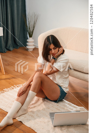Looking at laptop that is on the floor. Beautiful woman in casual clothes is at home in domestic room 128482584