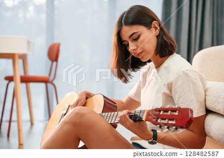 With classical guitar in hands. Beautiful woman in casual clothes is at home in domestic room 128482587