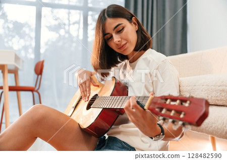 With classical guitar in hands. Beautiful woman in casual clothes is at home in domestic room With classical guitar in hands. Beautiful woman in casual clothes is at home in domestic room 128482590