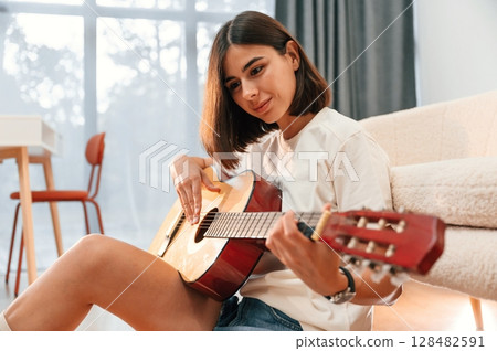 With classical guitar in hands. Beautiful woman in casual clothes is at home in domestic room With classical guitar in hands. Beautiful woman in casual clothes is at home in domestic room 128482591