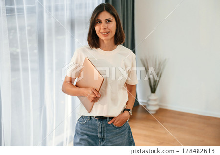 Standing and holding laptop in hands. Beautiful woman in casual clothes is at home in domestic room 128482613