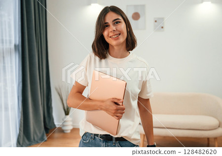 Smiling, holding laptop. Beautiful woman in casual clothes is at home in domestic room Smiling, holding laptop. Beautiful woman in casual clothes is at home in domestic room 128482620