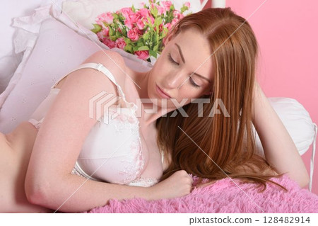 Beautiful redheaded model dressed in pink, lace lingerie, posing on her bed in an old house 128482914