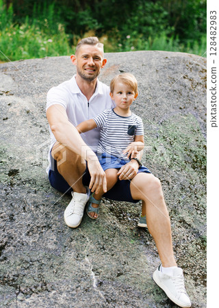 Smiling Father Sitting With Son On Large Rock In Nature Smiling Father Sitting With Son On Large Rock In Nature 128482983