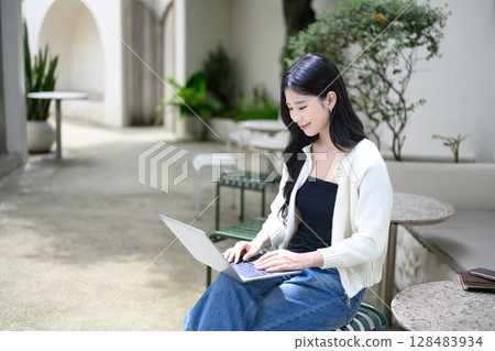Smiling woman working on laptop at a stylish stone table surrounded by plants Smiling woman working on laptop at a stylish stone table surrounded by plants 128483934