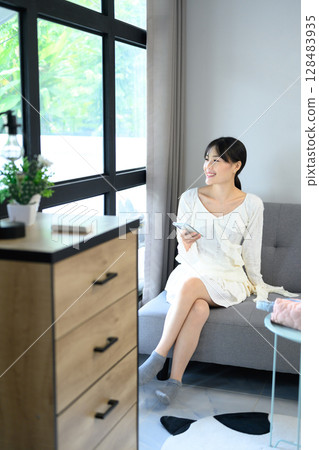 Asian woman sitting on a sofa, holding smartphone and looking out the window in a bright living room. Asian woman sitting on a sofa, holding smartphone and looking out the window in a bright living room. 128483935