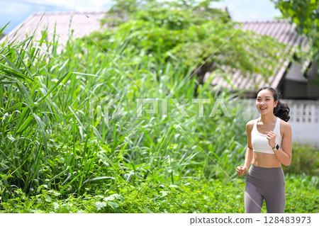 Smiling young woman jogging outdoors along a green path on a sunny day. Wellness concept 128483973