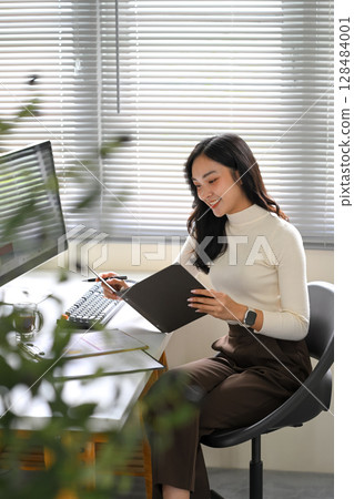 Young woman reviewing documents while working from home at a desk with desktop computer 128484001