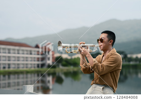 Confident musician playing trumpet with serene water and mountain views. Confident musician playing trumpet with serene water and mountain views. 128484008