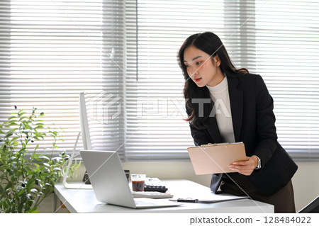 Young businesswoman holding a clipboard and analyzing data on a desktop computer 128484022