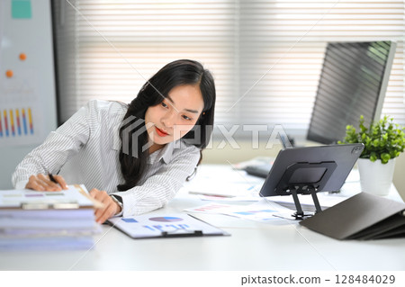 Office worker analyzing financial charts while reaching for stacked documents during work Office worker analyzing financial charts while reaching for stacked documents during work 128484029