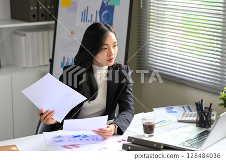 Asian businesswoman reviewing financial reports and data sheets at her desk 128484036