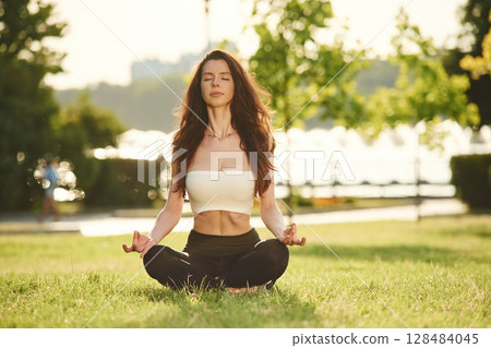 Meditating in lotus pose. Beautiful young woman in sportive clothes is in the park at sunny daytime 128484045