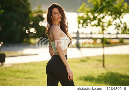 Standing, posing, showing body. Beautiful young woman in sportive clothes is in the park at sunny daytime 128484046